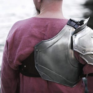 May include: A silver metal shoulder armor with a leather strap. The armor is worn over a pink shirt.