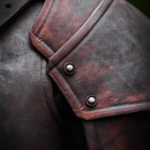 May include: Close-up of a brown leather shoulder armor with metal rivets. The armor has a distressed look and is made of thick leather.