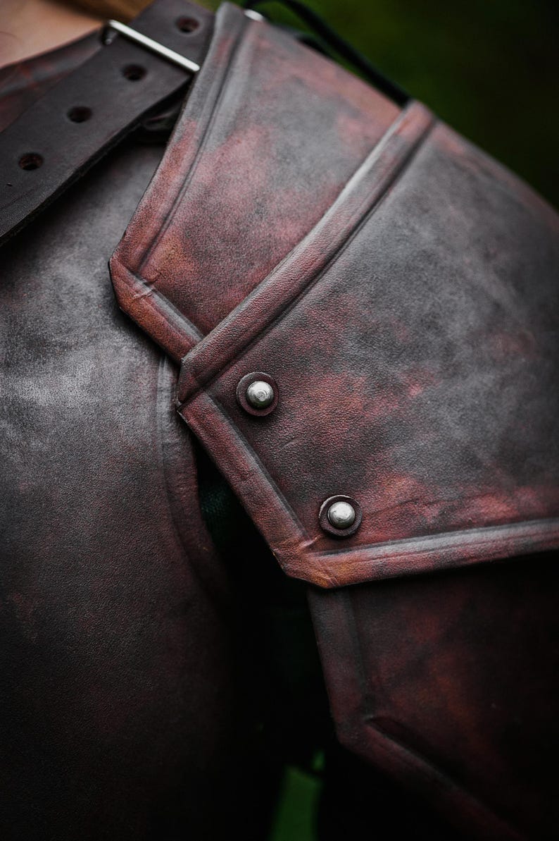 May include: Close-up of a brown leather shoulder armor with metal rivets. The armor has a distressed look and is made of thick leather.