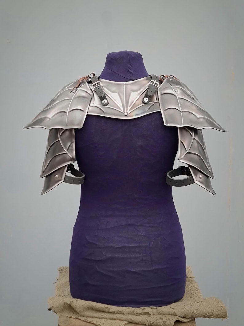 May include: Silver plated shoulder armor with a decorative design. The armor is made of metal and has straps for attaching it to the body.