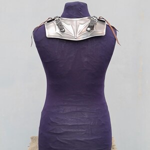 May include: Silver-colored metal shoulder armor with a decorative design. The armor has two adjustable leather straps with buckles.