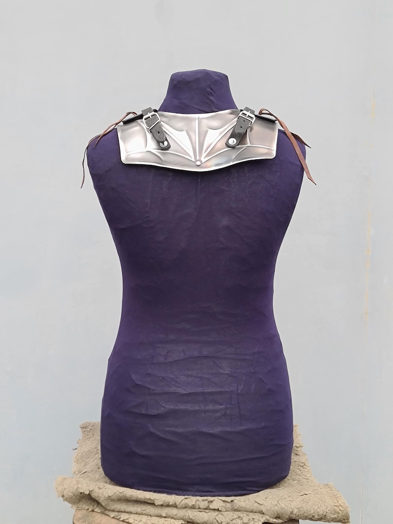 May include: Silver-colored metal shoulder armor with a decorative design. The armor has two adjustable leather straps with buckles.