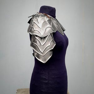May include: Silver plated shoulder armor with a decorative design. The armor is made of metal and has a layered look. It is displayed on a mannequin.