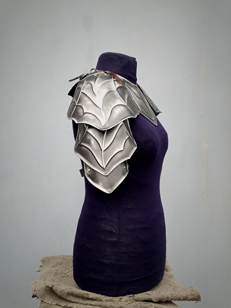 May include: Silver plated shoulder armor with a decorative design. The armor is made of metal and has a layered look. It is displayed on a mannequin.