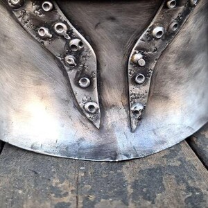 May include: A silver-colored metal helmet with riveted accents. The helmet has a curved design with decorative, pointed elements and a weathered appearance. The image shows the lower portion of the helmet, resting on a wooden surface.