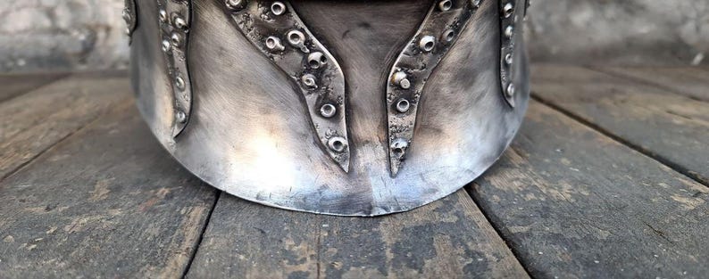 May include: A silver-colored metal helmet with riveted accents. The helmet has a curved design with decorative, pointed elements and a weathered appearance. The image shows the lower portion of the helmet, resting on a wooden surface.