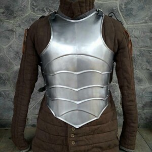 May include: A silver-colored metal breastplate with a decorative design. The breastplate is worn over a brown quilted jacket.