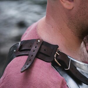 May include: A close-up of a brown leather strap with a metal buckle, attached to a piece of armor. The strap is worn over a pink shirt.