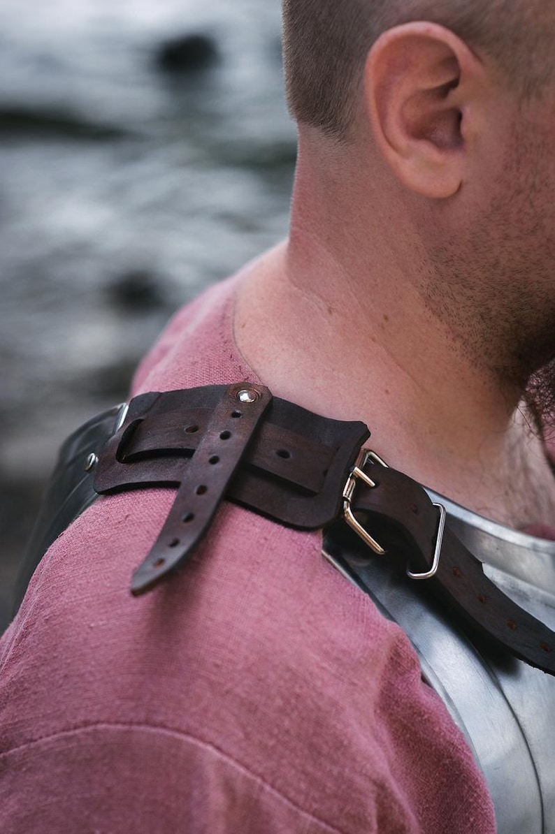 May include: A close-up of a brown leather strap with a metal buckle, attached to a piece of armor. The strap is worn over a pink shirt.