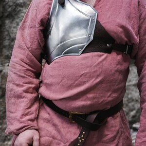 May include: A silver metal chest plate with a brown leather strap and buckle. The chest plate is worn over a pink linen shirt with a brown leather belt.