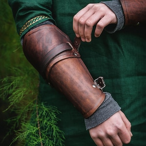 May include: A brown leather arm guard with a strap and buckle closure. The guard is worn on the left arm and is secured with a strap that wraps around the forearm.