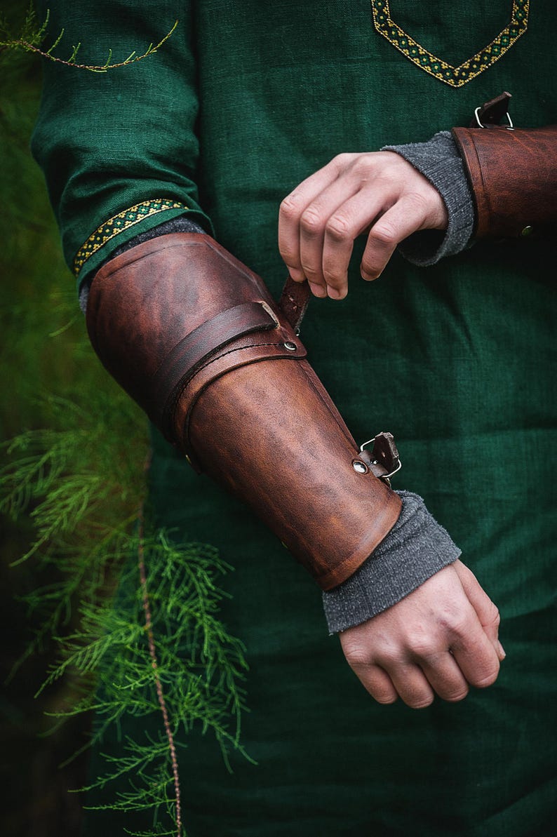 May include: A brown leather arm guard with a strap and buckle closure. The guard is worn on the left arm and is secured with a strap that wraps around the forearm.