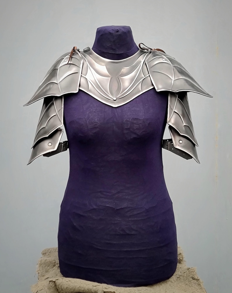 May include: Silver plated shoulder armor with a decorative design. The armor is displayed on a purple mannequin.