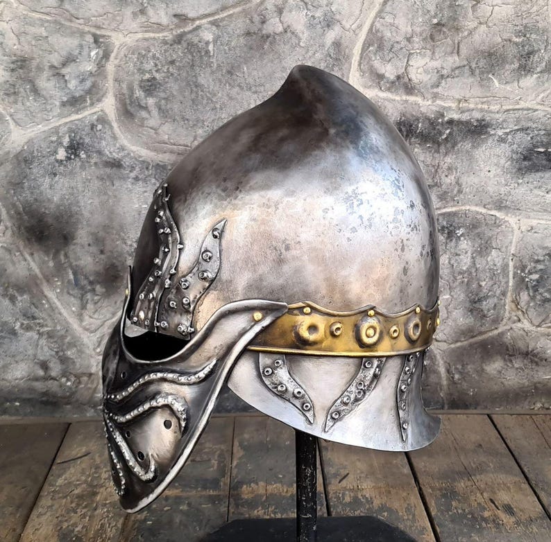 May include: A silver-colored medieval helmet with decorative accents. The helmet features a pointed top and a face guard with ornate detailing. A gold-colored band encircles the helmet near the base. The helmet is displayed on a black stand.