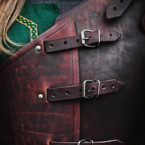 May include: A brown leather armor with three straps and buckles. The straps are secured with silver buckles. The armor is worn over a green and gray shirt.