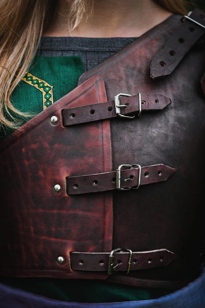 May include: A brown leather armor with three straps and buckles. The straps are secured with silver buckles. The armor is worn over a green and gray shirt.