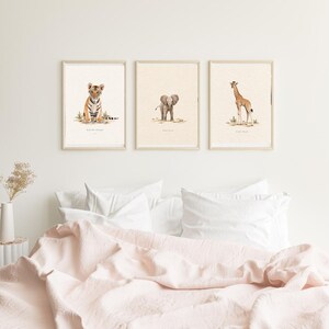 Puede incluir: Tres láminas decorativas enmarcadas con ilustraciones en acuarela de un cachorro de tigre, un elefante bebé y una jirafa joven. Cada animal aparece de pie o sentado sobre un fondo crema claro con detalles sutiles de suelo terroso.