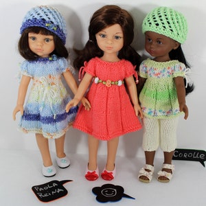 Op de afbeelding: Drie poppen die gebreide outfits dragen. De pop links draagt een blauw-wit gestreepte jurk met een blauwe hoed. De pop in het midden draagt een roze jurk met een witte bloemenriem. De pop rechts draagt een groen-wit gestreepte jurk met een groene hoed. De poppen staan op een wit oppervlak met een zwart schoolbord waarop "Corolle" staat en een ander bord waarop "Paola Reina" staat.