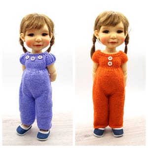 Puede incluir: Dos muñecas vestidas con monos de punto. Una muñeca lleva un mono azul bígaro con tres botones blancos, la otra un mono naranja con dos botones blancos. Ambas muñecas tienen coletas marrones y llevan zapatos azules.