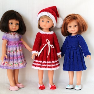 Peut inclure: Trois poupées portant des robes en tricot. La poupée de gauche porte une robe violette et rose. La poupée du milieu porte une robe rouge avec des bordures blanches et un bonnet de Père Noël blanc. La poupée de droite porte une robe bleue.