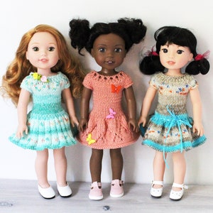 May include: Three dolls wearing knitted dresses. The doll on the left is wearing a teal and white dress with a floral embellishment. The doll in the middle is wearing a peach dress with butterfly embellishments. The doll on the right is wearing a brown and blue dress with a blue ribbon.
