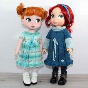 May include: Two dolls wearing knitted dresses. The doll on the left has red hair and is wearing a teal dress with a light blue ribbon. The doll on the right has red hair with a blue headband and is wearing a blue dress with a light blue ribbon.