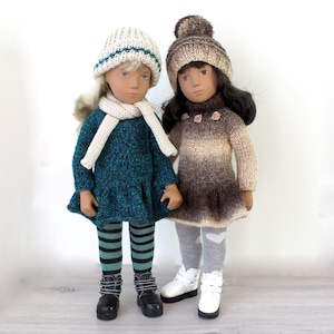 Könnte beinhalten: Zwei Puppen in gestrickten Winteroutfits. Die Puppe links trägt ein türkisfarbenes und schwarzes Strickkleid, einen weißen Schal und eine weiße Strickmütze mit einem blauen Streifen. Die Puppe rechts trägt ein braunes und beigefarbenes Ombre-Strickkleid, eine braune Strickmütze mit einem weißen Bommel und graue Strumpfhosen mit weißen Herzen.