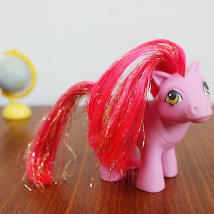 Op de afbeelding: Een roze speelgoedpony met een vloeiende rode en gouden manen en staart. De pony heeft gele ogen en staat op een houten oppervlak. Een klein globe-speelgoed staat op de achtergrond.