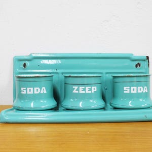 Op de afbeelding: Een turquoise geëmailleerd wandrek met drie bijpassende cilindrische bakjes. Twee bakjes zijn voorzien van de tekst SODA en één van de tekst ZEEP in witte blokletters. Het metalen rek heeft een rechthoekige achterplaat met montagegaten.