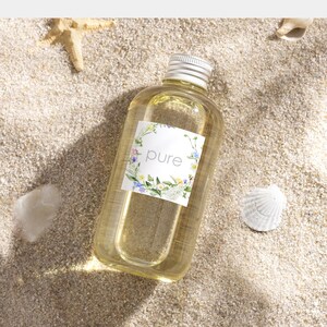 May include: A clear glass bottle with a silver cap filled with a light yellow liquid. The bottle has a white label with a floral wreath and the word "pure" in the center.
