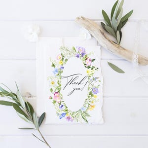 May include: A white card with a watercolor wreath of flowers in shades of pink, purple, yellow, and green. The wreath surrounds a white oval with the text "Thank you!" in black calligraphy.