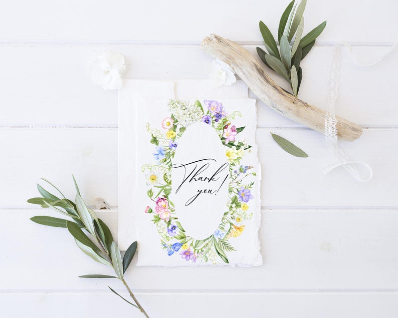 May include: A white card with a watercolor wreath of flowers in shades of pink, purple, yellow, and green. The wreath surrounds a white oval with the text "Thank you!" in black calligraphy.