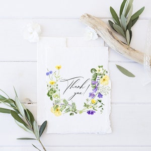 May include: A white card with a watercolor floral wreath in shades of purple, yellow, and green. The wreath surrounds the text "Thank you!" in black calligraphy.