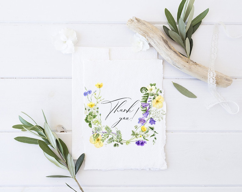 May include: A white card with a watercolor floral wreath in shades of purple, yellow, and green. The wreath surrounds the text "Thank you!" in black calligraphy.
