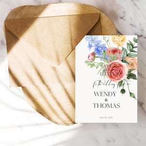 Op de afbeelding: Een trouwuitnodigingskaart met een bloemenontwerp, met roze, perzik en blauwe bloemen. De kaart leest "Save the Date" en de namen "Wendy & Thomas" met de datum 18 juli 2025. Een bruine envelop is inbegrepen.