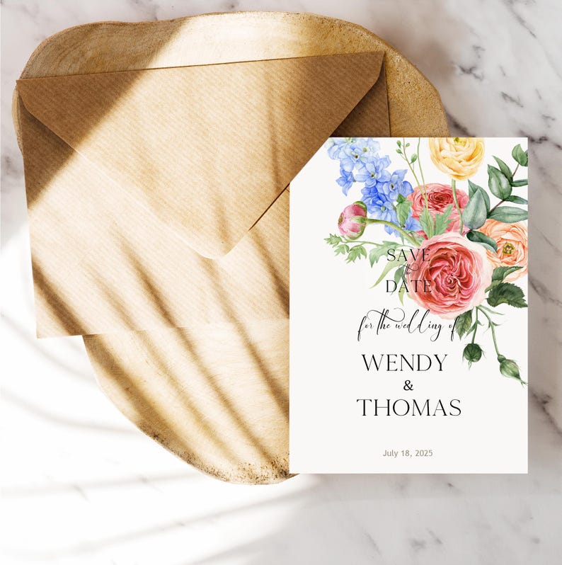 Op de afbeelding: Een trouwuitnodigingskaart met een bloemenontwerp, met roze, perzik en blauwe bloemen. De kaart leest "Save the Date" en de namen "Wendy & Thomas" met de datum 18 juli 2025. Een bruine envelop is inbegrepen.