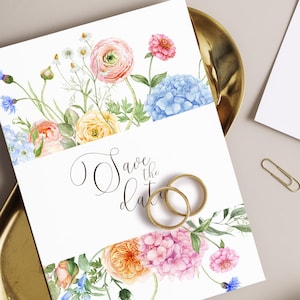 Op de afbeelding: Een witte huwelijksuitnodiging met bloemenranden en de tekst "Save the date". Het ontwerp toont roze, oranje, blauwe en gele bloemen, met twee gouden ringen.