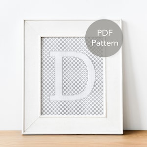 May include: A white framed picture with a black and white cross-stitch pattern of the letter "D" inside. The frame has a white mat and the text "PDF Pattern" is written in a circle in the top right corner.