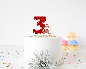 May include: A white cake with red number 3 and a sock monkey topper. The cake is decorated with colorful sprinkles. There are three cupcakes in the background.