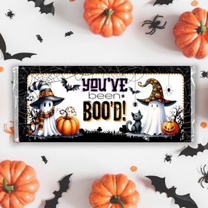 May include: A Halloween-themed chocolate bar wrapper with a ghost wearing a witch hat and holding a pumpkin. The wrapper has a black and white spiderweb design and the text "You've been BOO'D!"