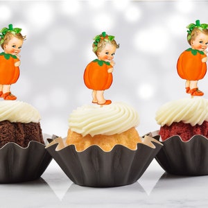 May include: Three cupcakes with black cupcake liners and white frosting. Each cupcake is topped with a pumpkin-shaped cake topper featuring a baby dressed in a pumpkin costume.