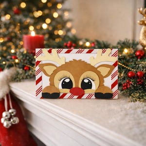 May include: A rectangular greeting card featuring a cartoon reindeer with large eyes, long eyelashes, and a red nose. The reindeer is framed by a white border with diagonal red and white candy cane stripes. The card is made of layered paper cutouts.