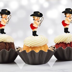 May include: Three cupcakes with black cupcake liners and white frosting. Each cupcake has a topper of a baby girl wearing a black cowboy hat and red ruffled diaper, holding a lasso.