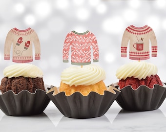 May include: Three cupcakes with white frosting, each topped with a paper cutout of a sweater. The sweaters have different designs, including mittens, leaves, and a mug. The cupcakes are in black paper cups.