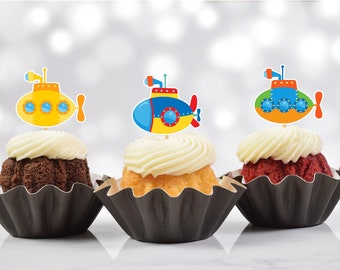 May include: Three cupcakes with white frosting and submarine-shaped toppers. The cupcakes are in black paper cups, with one chocolate, one vanilla, and one red velvet. The submarine toppers are yellow, blue, and green.