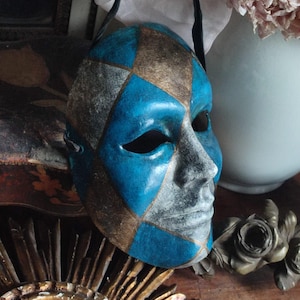 May include: A decorative mask with a harlequin pattern in blue, gold, and silver. The mask has a black strap and is likely made of papier-mâché or a similar material. The mask is a theatrical or costume item.