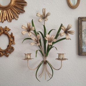 May include: Cream-colored metal wall sconce with a floral design, featuring stylized flowers and green leaves. The sconce has two candle holders and is mounted on a white wall, alongside other decorative frames and mirrors.