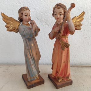 Może przedstawiać: Dwie figurki aniołów w stylu vintage. Jeden anioł gra na flecie, drugi na lutni. Oba mają złote skrzydła i ubrane są w długie szaty. Figurki pomalowane są w stonowanych odcieniach niebieskiego, brzoskwiniowego i złotego i stoją na małych drewnianych podstawach.