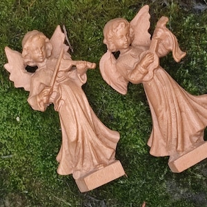 Può includere: Due statuette di angeli in legno, una che suona il violino e l'altra il liuto. Gli angeli sono marrone chiaro con ali dettagliate e vesti fluenti. Si ergono su basi rettangolari, su uno sfondo di muschio verde.