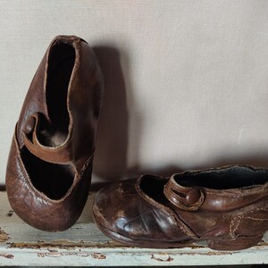 Könnte beinhalten: Ein Paar antike braune Mary Jane-Schuhe aus Leder. Die Schuhe haben eine runde Spitze, einen Riemen über dem Oberteil mit Knopfverschluss und einen niedrigen Absatz. Das Leder weist Gebrauchsspuren und Alter auf, mit sichtbaren Falten und Kratzern.